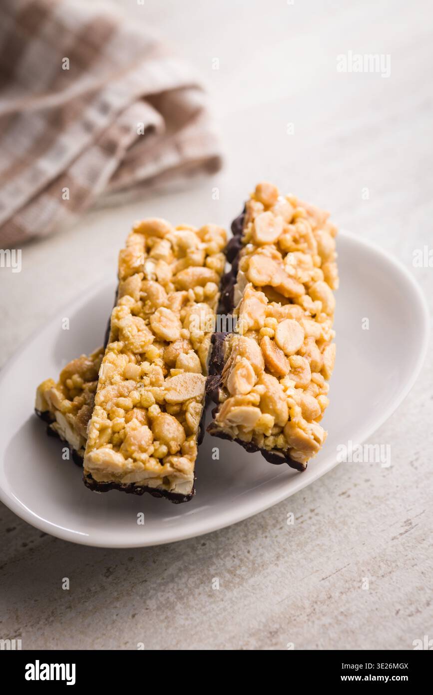 Barres de granola aux arachides empilées avec une base enrobée de chocolat sur une assiette blanche, serties sur un chiffon rayé. Croustillant, snack sucré idéal pour le petit déjeuner, Lunchbox Banque D'Images
