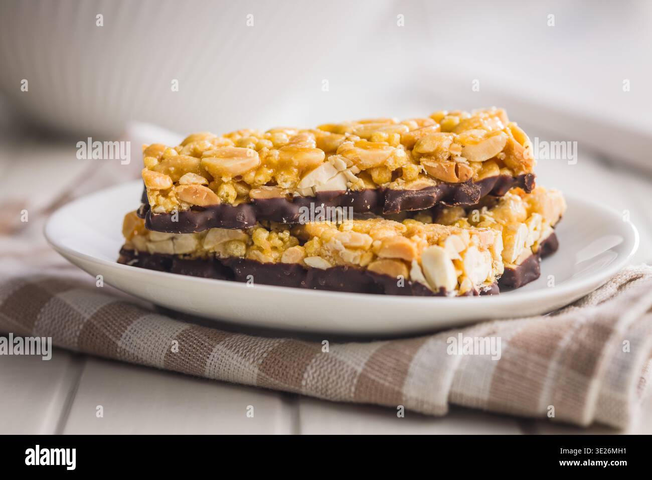 Barres de granola aux arachides empilées avec une base enrobée de chocolat sur une assiette blanche, serties sur un chiffon rayé. Croustillant, snack sucré idéal pour le petit déjeuner, Lunchbox Banque D'Images