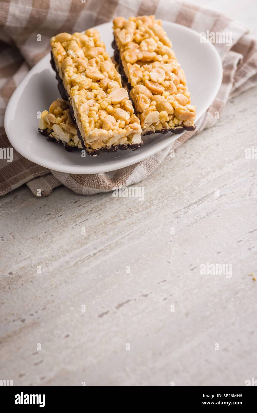 Barres de granola aux arachides empilées avec une base enrobée de chocolat sur une assiette blanche, serties sur un chiffon rayé. Croustillant, snack sucré idéal pour le petit déjeuner, Lunchbox Banque D'Images