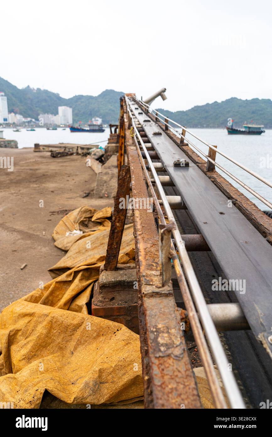 Un vieux système de bande transporteuse rouillée est installé sur une jetée en béton surplombant une baie avec des navires et des collines en arrière-plan Banque D'Images