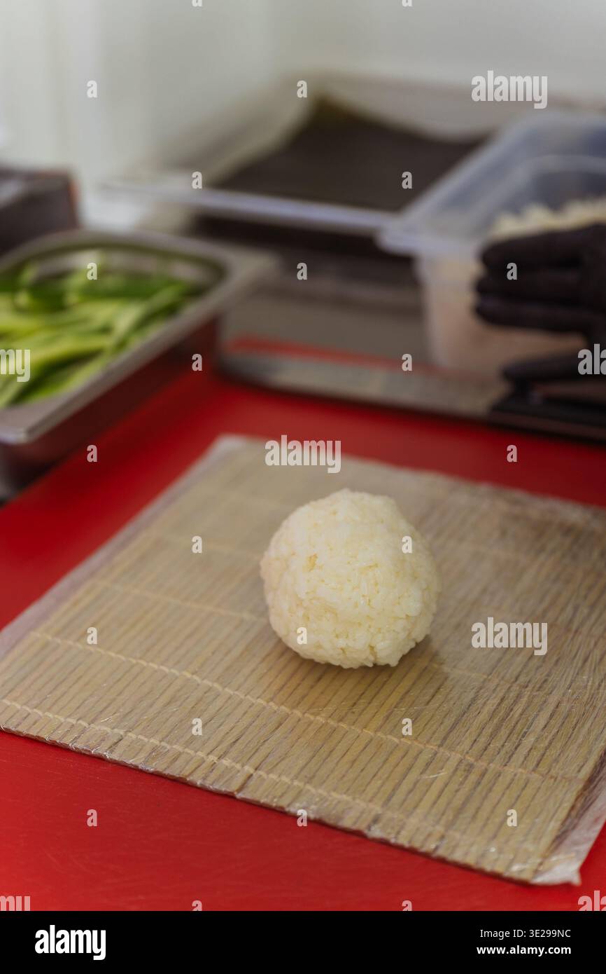 Boule de riz simple sur tapis de bambou, reposant sur une planche à découper rouge, plateaux d'ingrédients flous en arrière-plan Banque D'Images