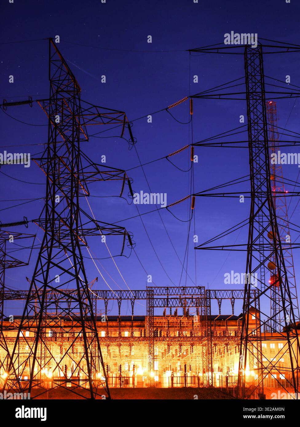 Vue nocturne d'une sous-station électrique avec des structures éclairées, des tours de transmission et des câbles sous un ciel bleu foncé pendant une longue exposition. Banque D'Images