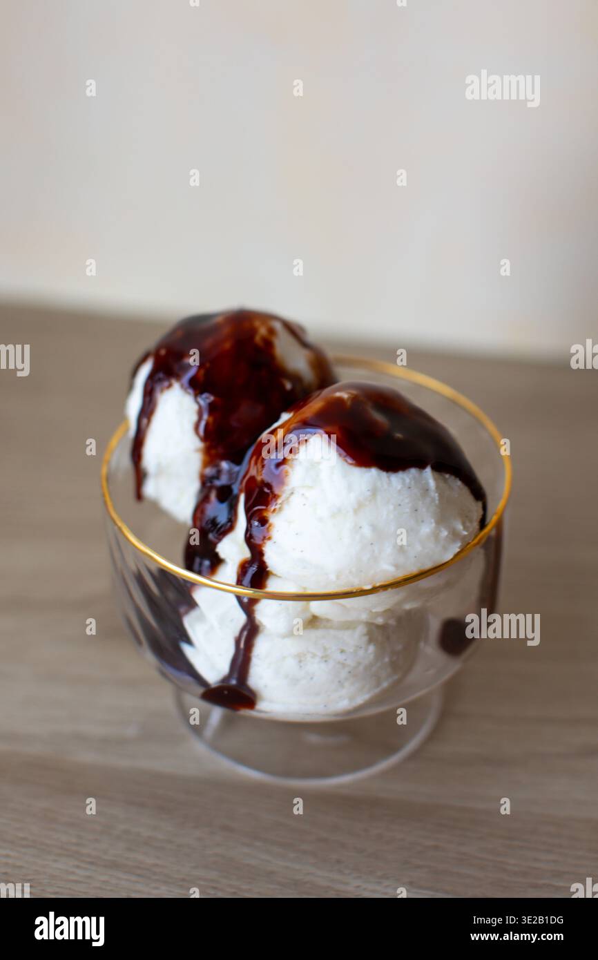 des boules de crème glacée aux haricots de vanille servies dans un bol à dessert en verre transparent avec un bord doré. Le dessert est arrosé de sirop de chocolat noir riche ou de fudge Banque D'Images