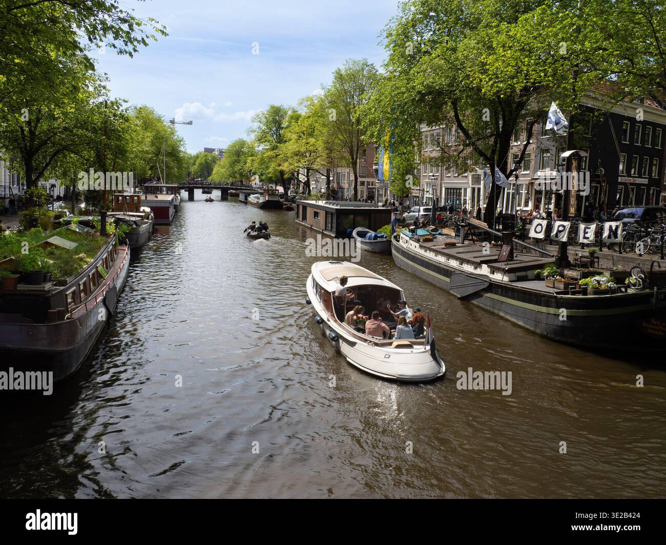Une scène sereine sur le canal d'Amsterdam, en Hollande, avec un bateau blanc naviguant devant des bâtiments historiques en briques et une végétation luxuriante. Banque D'Images