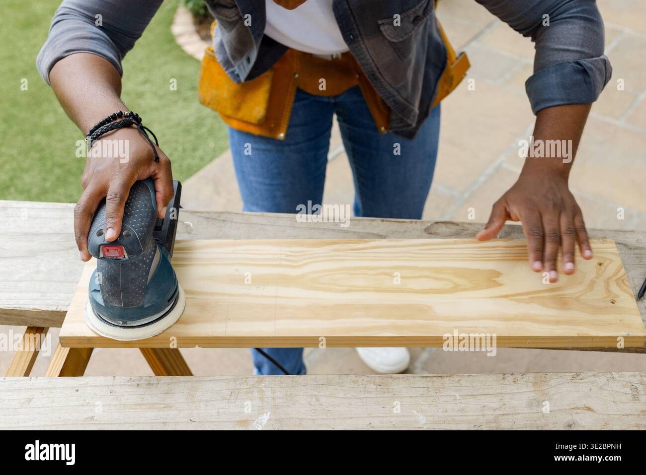 Homme afro-américain ponçant la planche de pin sur les sawhorses d'arrière-cour à l'aide d'une ponceuse orbitale, chemise en denim. Extérieur, patio, travail du bois, établi, bois, artisanat Banque D'Images