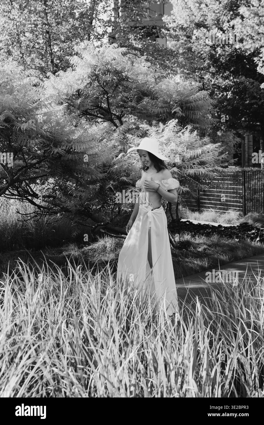 Une femme se promène dans un jardin rempli de hautes herbes et de verdure elle porte un chapeau et une robe légère tout en profitant du plein air en plein soleil Banque D'Images