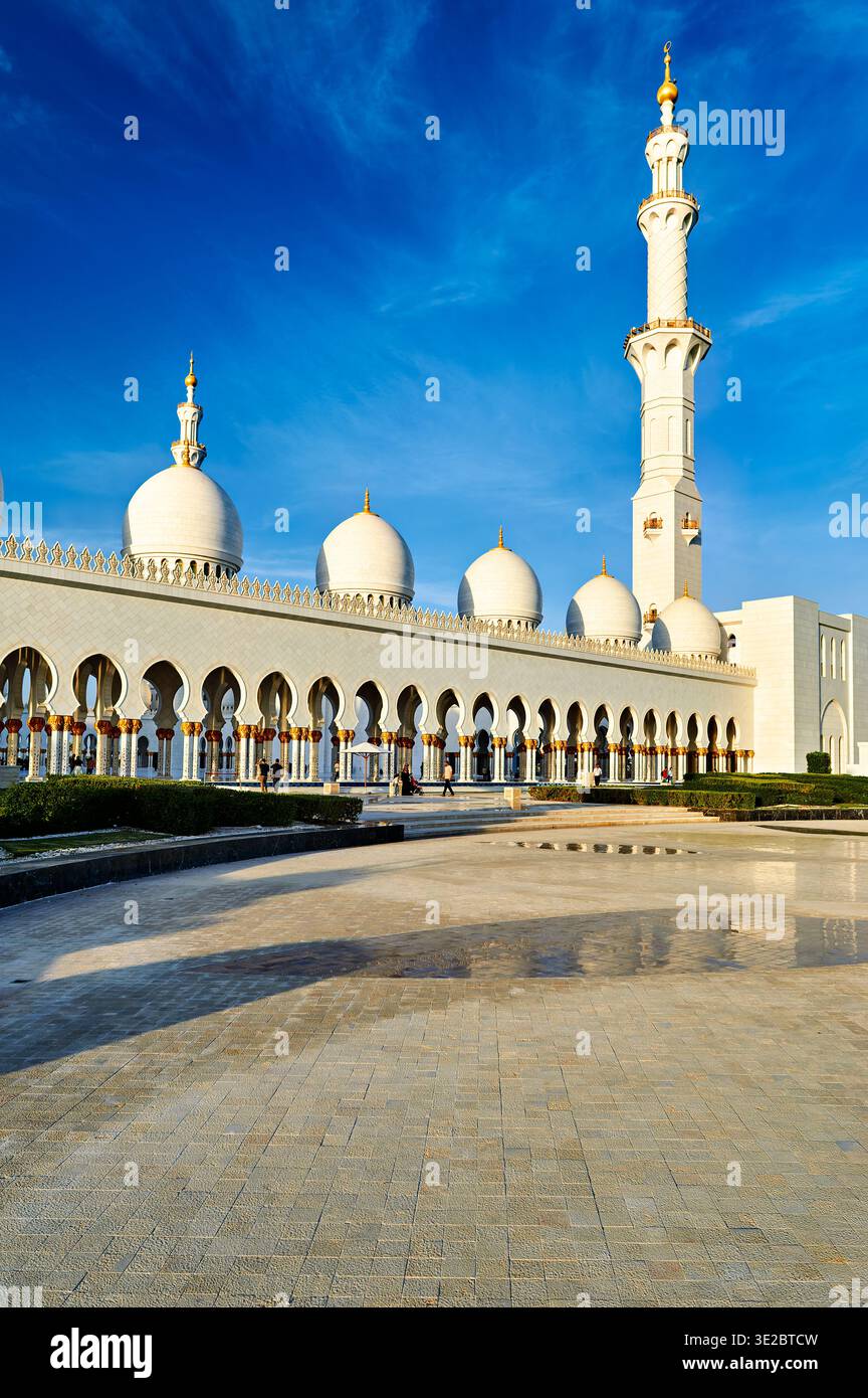 La mosquée d'Abu Dhabi présente ses grands dômes et son grand minaret contre un ciel dégagé. Les visiteurs se promènent dans la cour. Banque D'Images