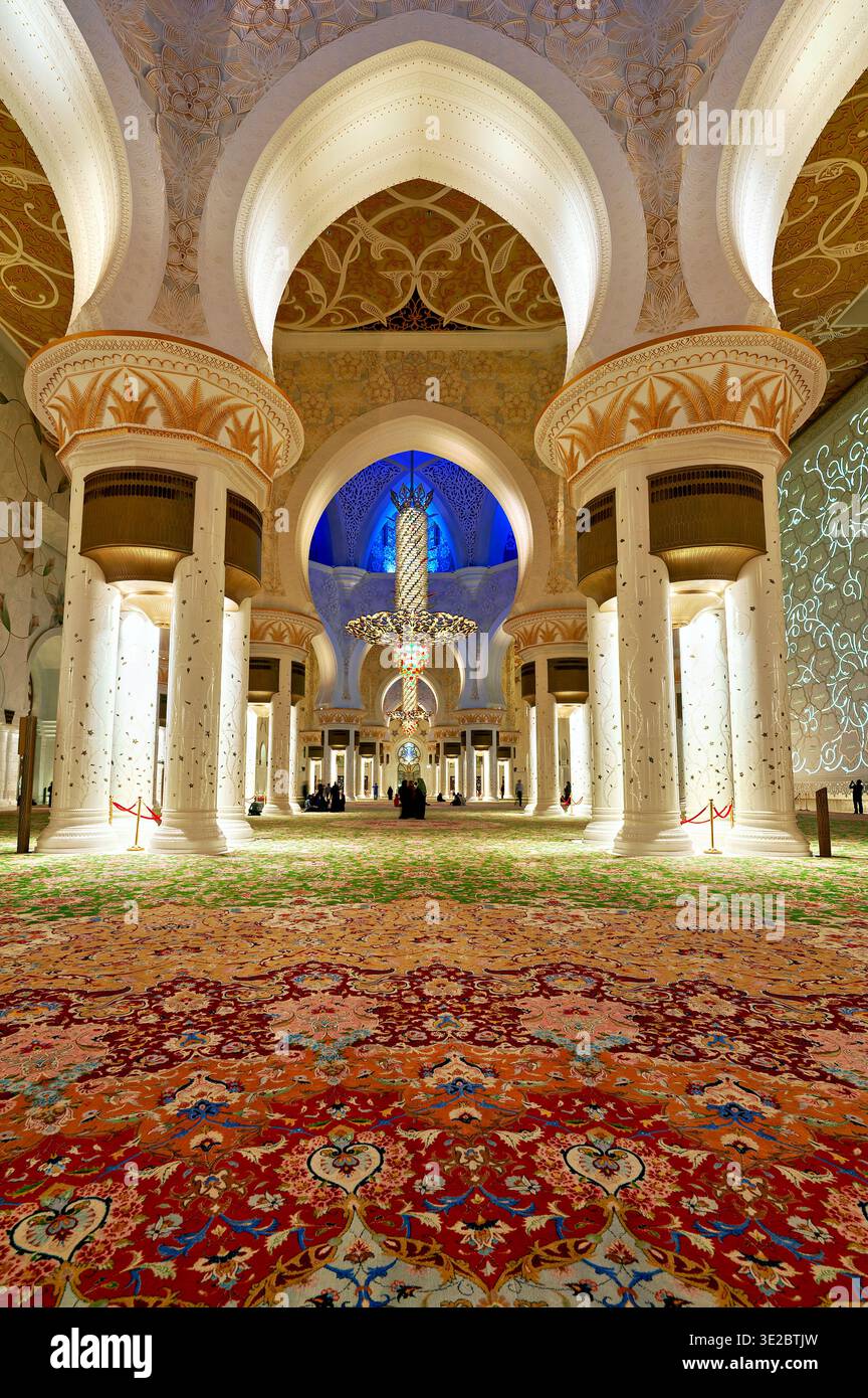 Les visiteurs se promènent dans le hall principal de la Grande Mosquée Sheikh Zayed à Abu Dhabi, entouré de grandes arches et de lumières décoratives. Banque D'Images