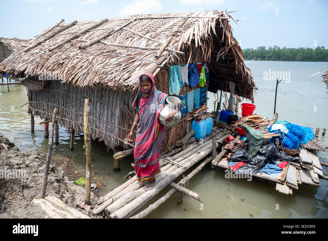 Khulna, Bangladesh - 12 juillet 2025 : vue d'une femme marchant sur une planche de bois portant un pot près de maisons sur pilotis aux toits de chaume, contrastant avec th Banque D'Images