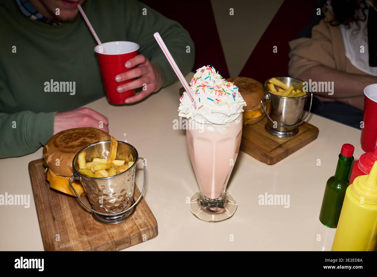 Amis inconnus assis à table buvant des milkshakes et mangeant des cheeseburgers avec des frites, mains visibles tenant des tasses rouges, milkshake avec de la crème fouettée et des saupoudres au premier plan Banque D'Images