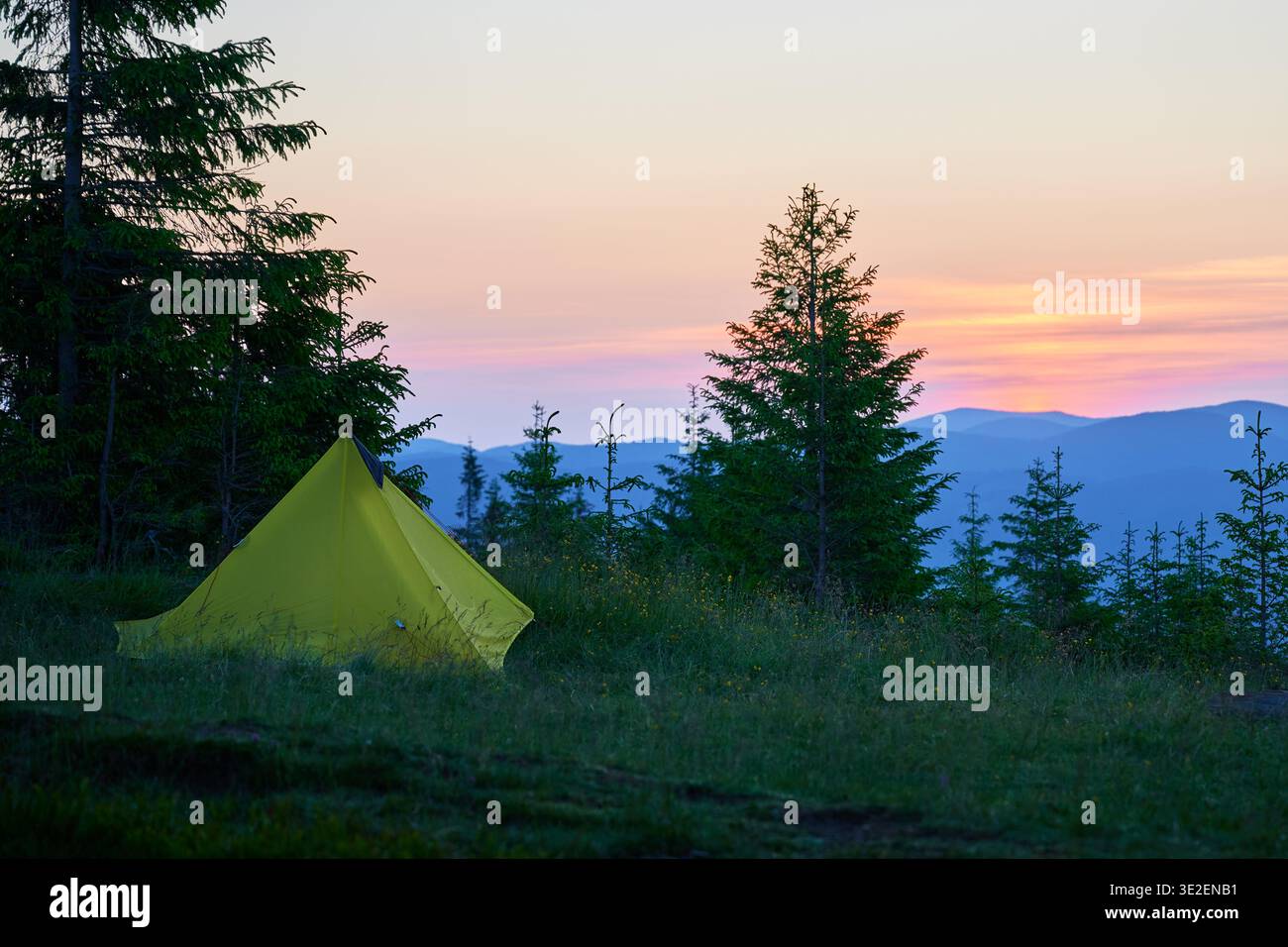 Camping serein avec tente jaune au milieu d'une verdure luxuriante, de pins, rétroéclairé par des teintes douces de lever de soleil. Les transitions du ciel des oranges chaudes aux bleus froids, les montagnes lointaines créent une toile de fond paisible. Banque D'Images