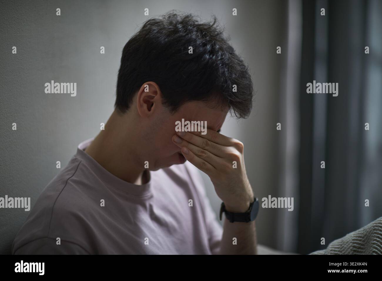 Jeune homme caucasien adulte assis contre le mur couvrant le visage avec la main montrant l'expression stressée, cheveux bruns courts visibles, portant montre-bracelet, transmettant l'émotion Banque D'Images