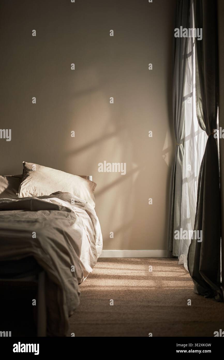 Vertical de l'intérieur de la chambre vide montrant un lit non fait avec la lumière du soleil qui coule à travers la fenêtre, projetant des ombres sur le mur et le sol, évoquant un sentiment de solitude et une atmosphère émotionnelle calme Banque D'Images