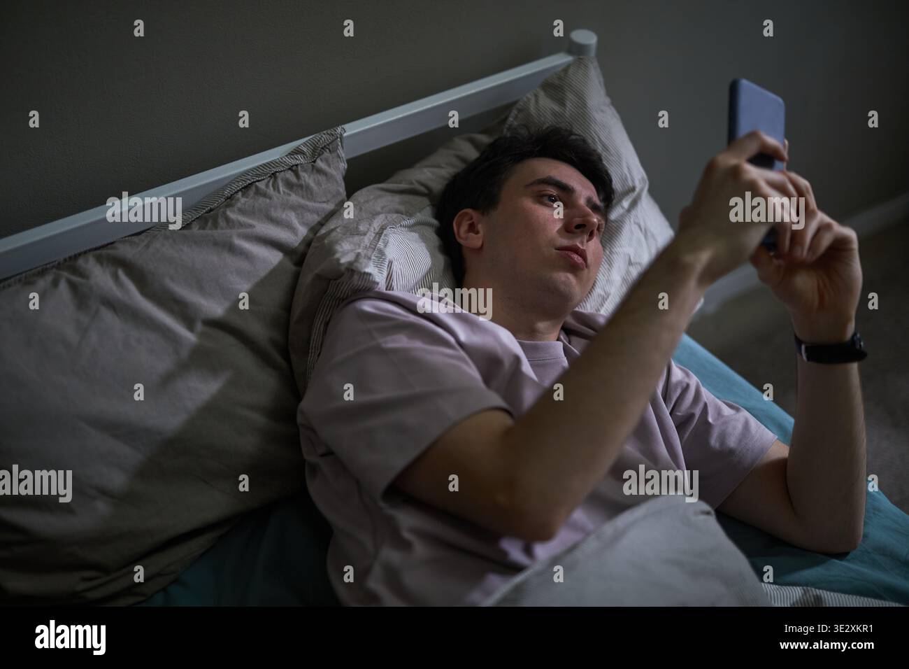 Jeune homme caucasien adulte allongé dans le lit tenant un smartphone au-dessus du visage, affichant une expression neutre ou contemplative tout en utilisant l'appareil, la tête reposant sur un oreiller, s'engageant avec l'écran Banque D'Images