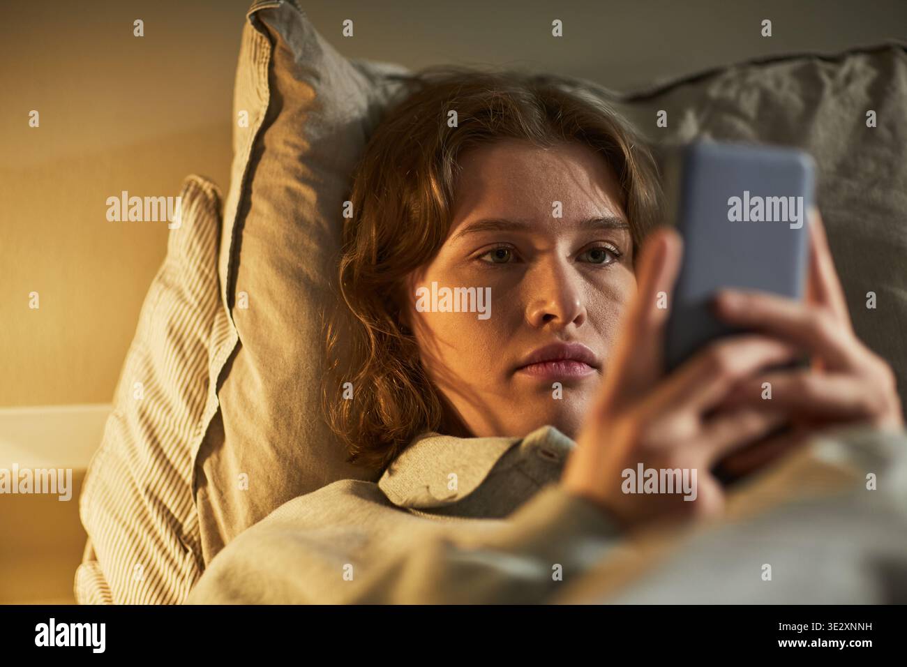 Jeune femme caucasienne adulte allongée dans le lit tenant un smartphone, regardant l'écran avec une expression faciale sérieuse, transmettant l'émotion à travers les yeux focalisés et la posture détendue Banque D'Images