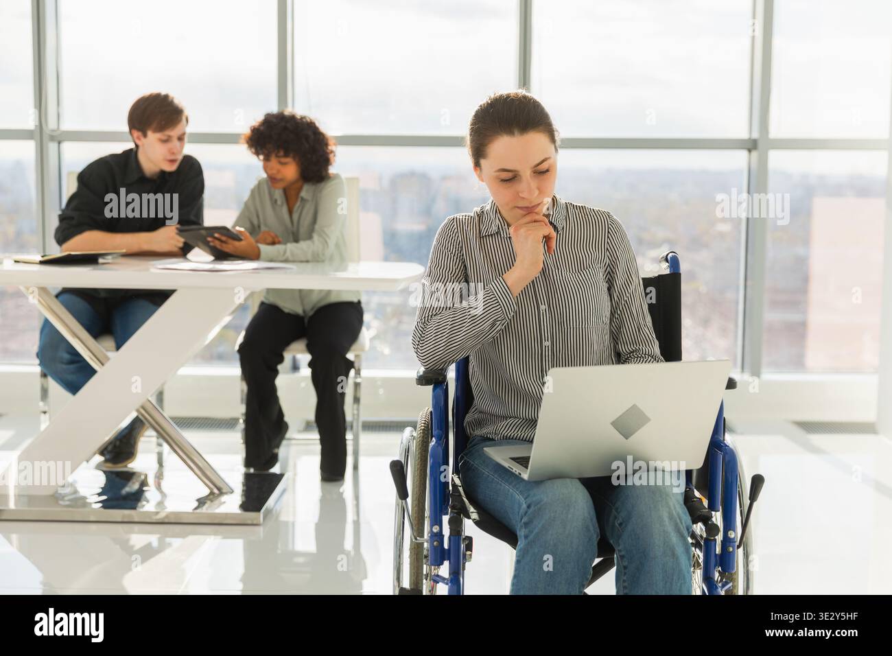 Environnement accessible. Femme en chaise pour les personnes handicapées travaillant avec un ordinateur portable dans un bureau moderne. Groupe multiracial diversifié de collègues travaillant ensemble. Jeune équipe professionnelle Banque D'Images