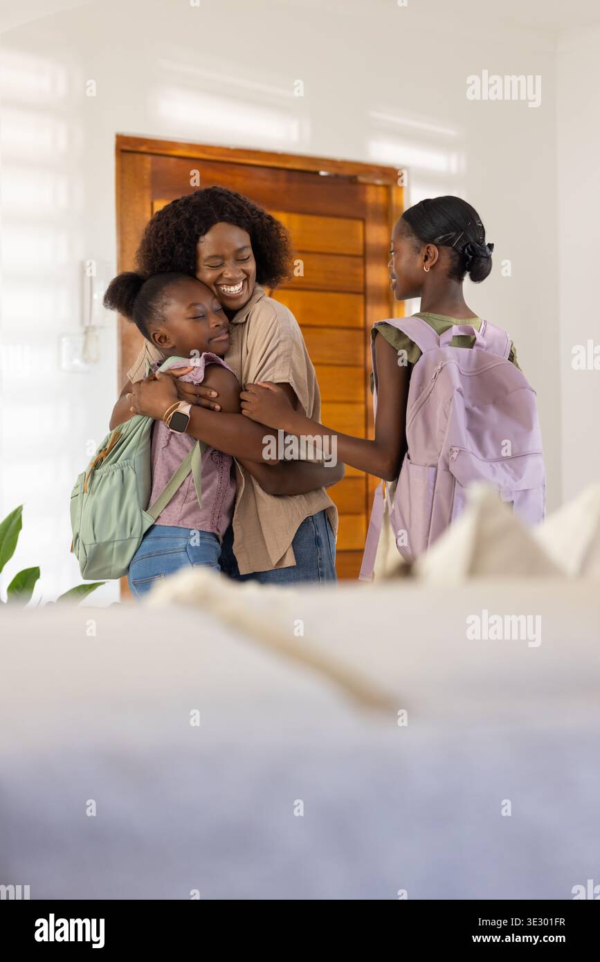 Mère africaine embrassant deux filles à l'entrée de la maison, tenant des sacs à dos près de la porte en bois Banque D'Images