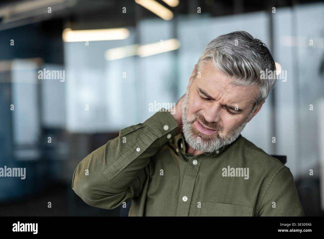 Homme d'affaires mûr avec les cheveux gris et la barbe frottant son cou dans un bureau moderne, grimacant de douleurs cervicales et une mauvaise posture tout en travaillant à un ordinateur, stressé et fatigué Banque D'Images