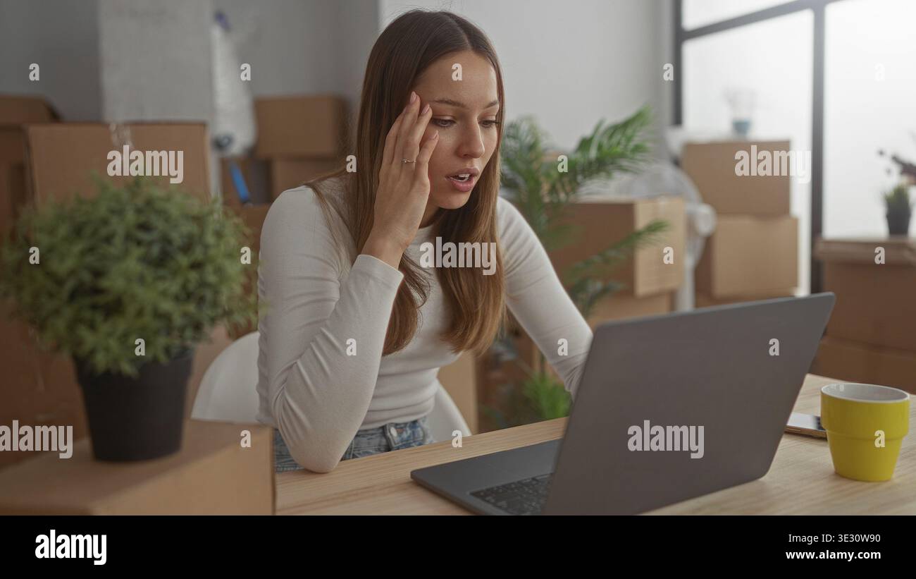 Femme avec la main sur la joue regardant l'ordinateur portable dans le bâtiment entouré de boîtes en mouvement emballées, plante, et tasse sur la table ; stress de jour en mouvement. Banque D'Images