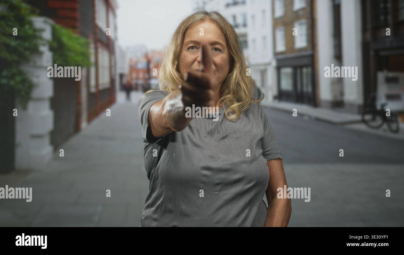 Femme âgée aux cheveux blonds pointe doigt directement à la caméra sur une rue urbaine bordée de bâtiments et de vélos ; confiance. Banque D'Images