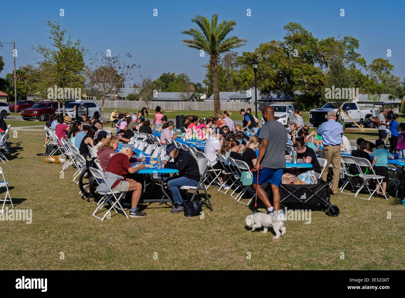 Lynn Haven, Floride, États-Unis. 20 mars 2026. Des dizaines de membres de la communauté se rassemblent autour de longues tables pliantes bleues à Sheffield Park pour peindre sur des toiles fournies gratuitement lors de l’événement Art in the Park parrainé par la ville. Les familles, les amis et les résidents locaux de tous âges et de tous niveaux de compétence profitent d’un après-midi ensoleillé exprimant leur créativité ensemble dans un magnifique cadre extérieur. Au premier plan, un homme en chemise grise promène un petit chien blanc devant la scène animée tandis que les participants se concentrent sur la création de leur prochain grand chef-d'œuvre. Crédit : Tosh Persons / Alamy Live News Banque D'Images