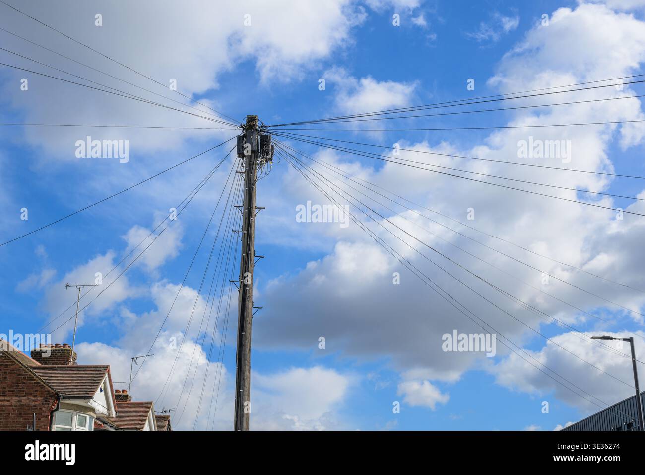 Poteau télégraphique en bois avec plusieurs fils électriques aériens s'étendant vers l'extérieur contre un ciel bleu avec des nuages dans un cadre de rue résidentiel. Banque D'Images