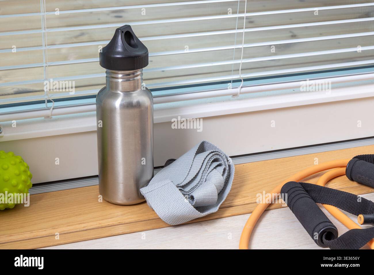 Bouteille d'eau en métal avec sangle de yoga roulée, bande de résistance et balle de massage placée sur le plancher en bois près des stores. Salle de fitness intérieure lumineuse Banque D'Images