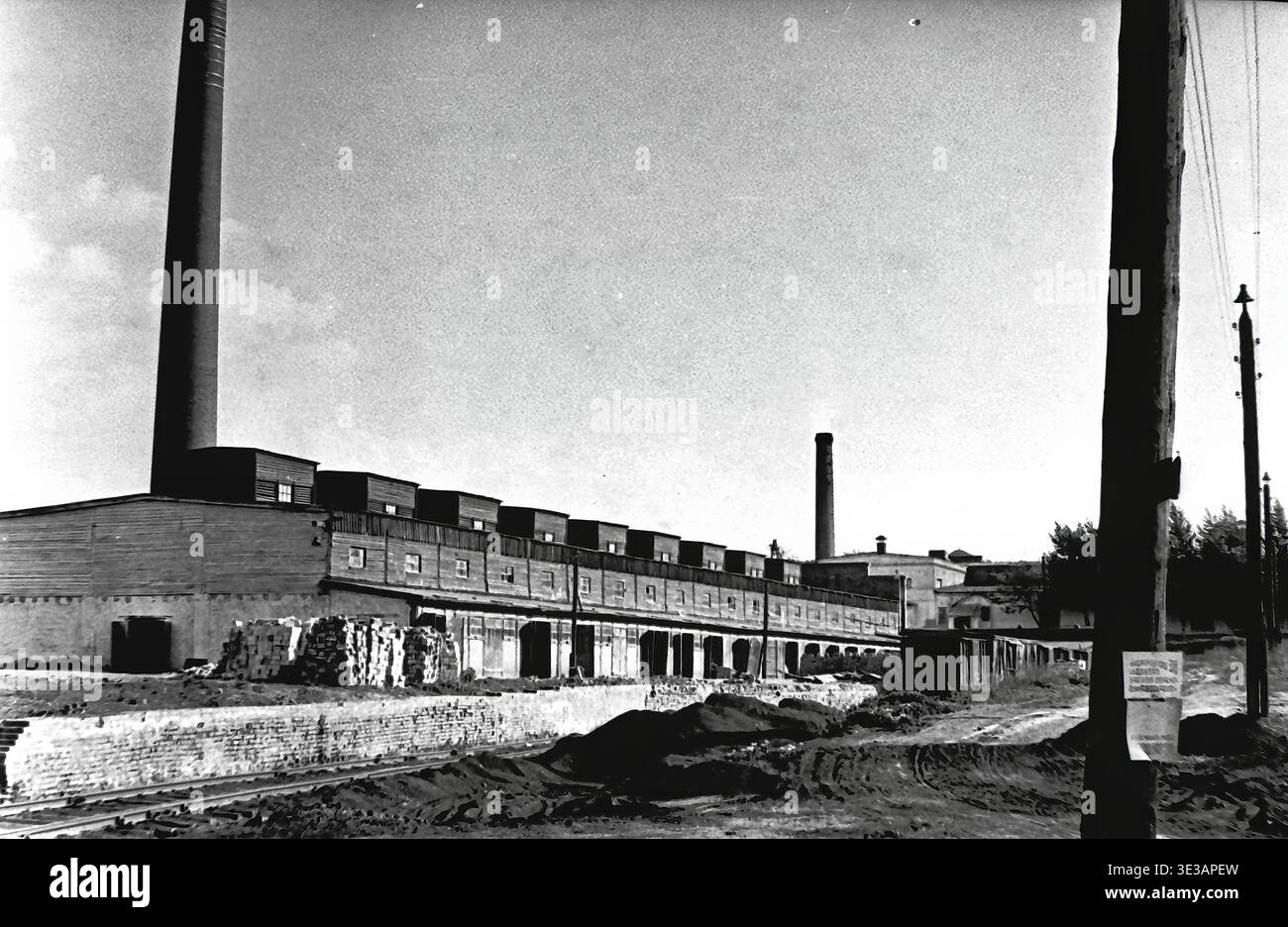 Sloviansk, années 1950-1960 Usine AIZ (usine d'armature-isolateur). Photographie d'archive vintage montrant le vaste territoire industriel avec un long bâtiment de fabrication et un grand smokestack. Des piles de briques et de matériaux de construction se trouvent au premier plan sur le sol brut, illustrant l'expansion active et le fonctionnement continu du complexe de l'industrie lourde soviétique. Cet impressionnant cliché en noir et blanc documente le patrimoine architectural authentique, préservant l'époque du Donbass paisible. Banque D'Images