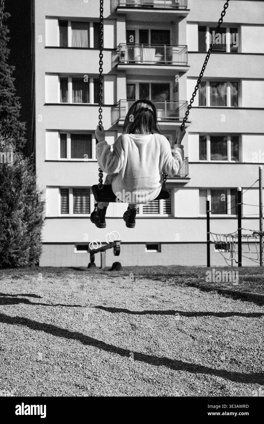 Photo en noir et blanc de l'enfant assis sur la balançoire dans l'aire de jeux urbaine avec bâtiment résidentiel en arrière-plan. Scène de style de vie quotidienne capturant l'enfant Banque D'Images