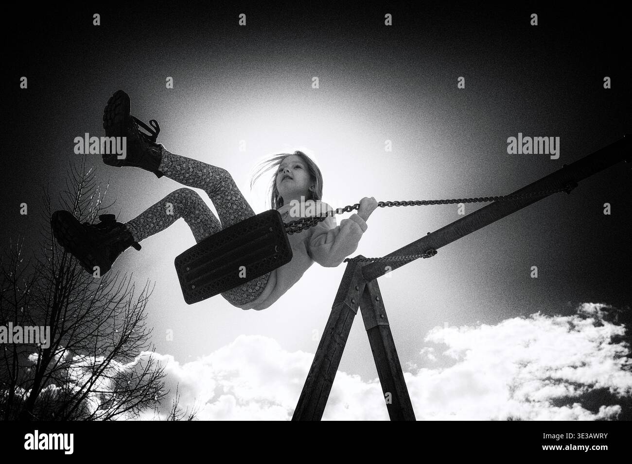 Photo en noir et blanc d'un enfant se balançant haut contre un ciel lumineux avec une forte lumière du soleil. Scène dynamique et émotionnelle symbolisant la liberté, la joie et le carefre Banque D'Images