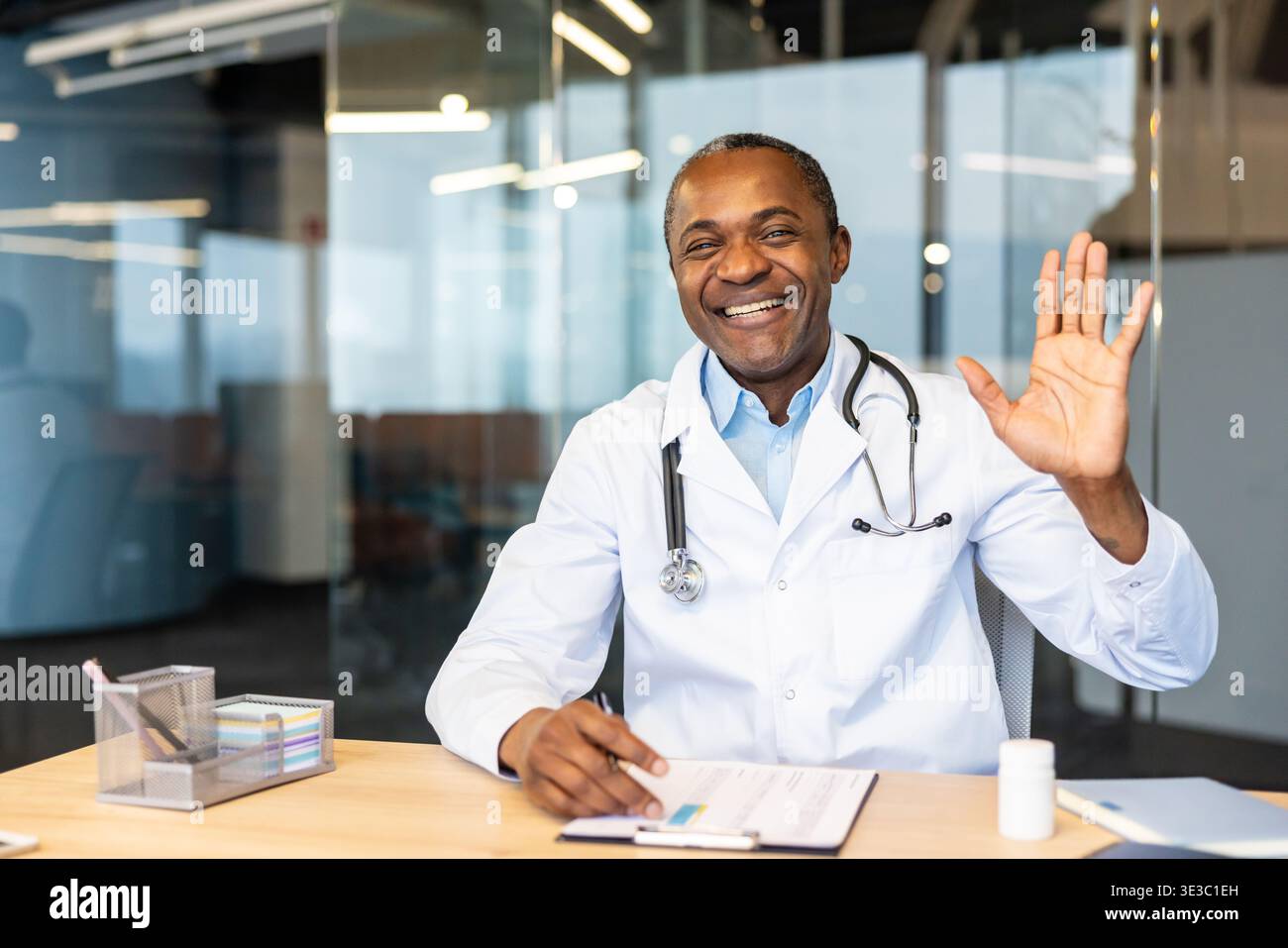 Médecin afro-américain portant une blouse de laboratoire blanche et un stéthoscope, souriant chaleureusement et agitant la main tout en étant assis à un bureau dans un cabinet médical moderne, fournissant des soins virtuels Banque D'Images