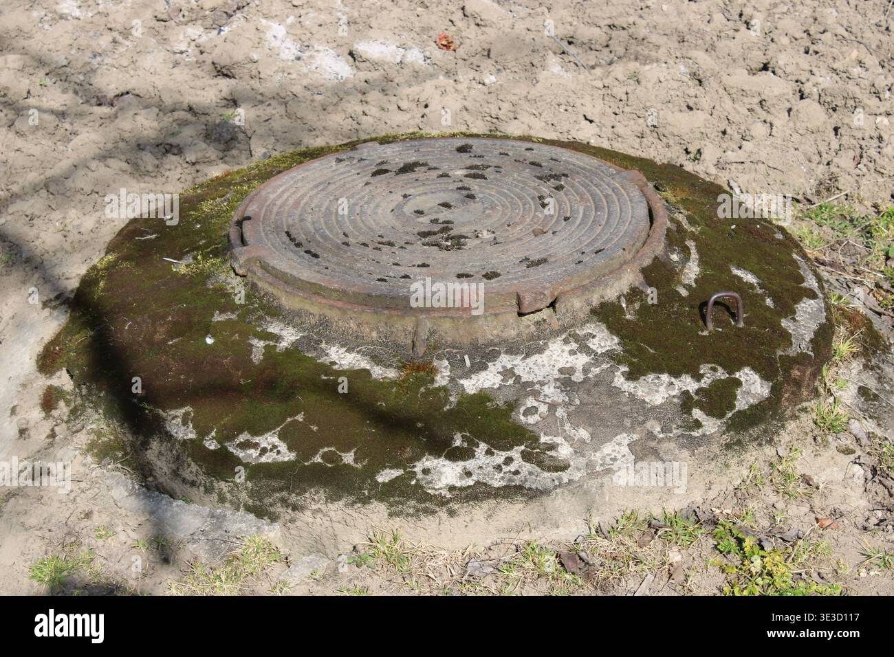 Un couvercle de trou d'homme repose sur une parcelle de terre et d'herbe. La surface montre une certaine usure et la croissance de mousse. La lumière du soleil brille, soulignant sa forme ronde aga Banque D'Images