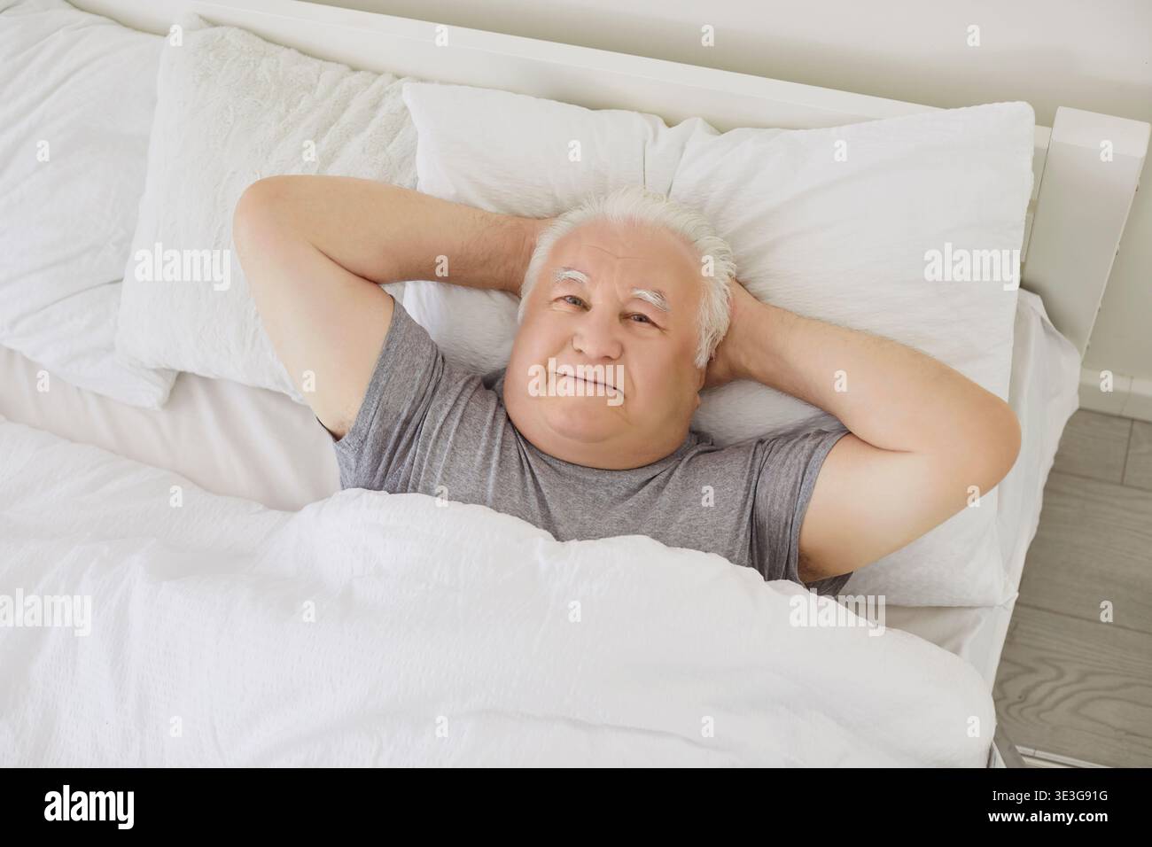 Heureux homme âgé retraité souriant couché avec les mains derrière la tête dans le lit le matin Banque D'Images