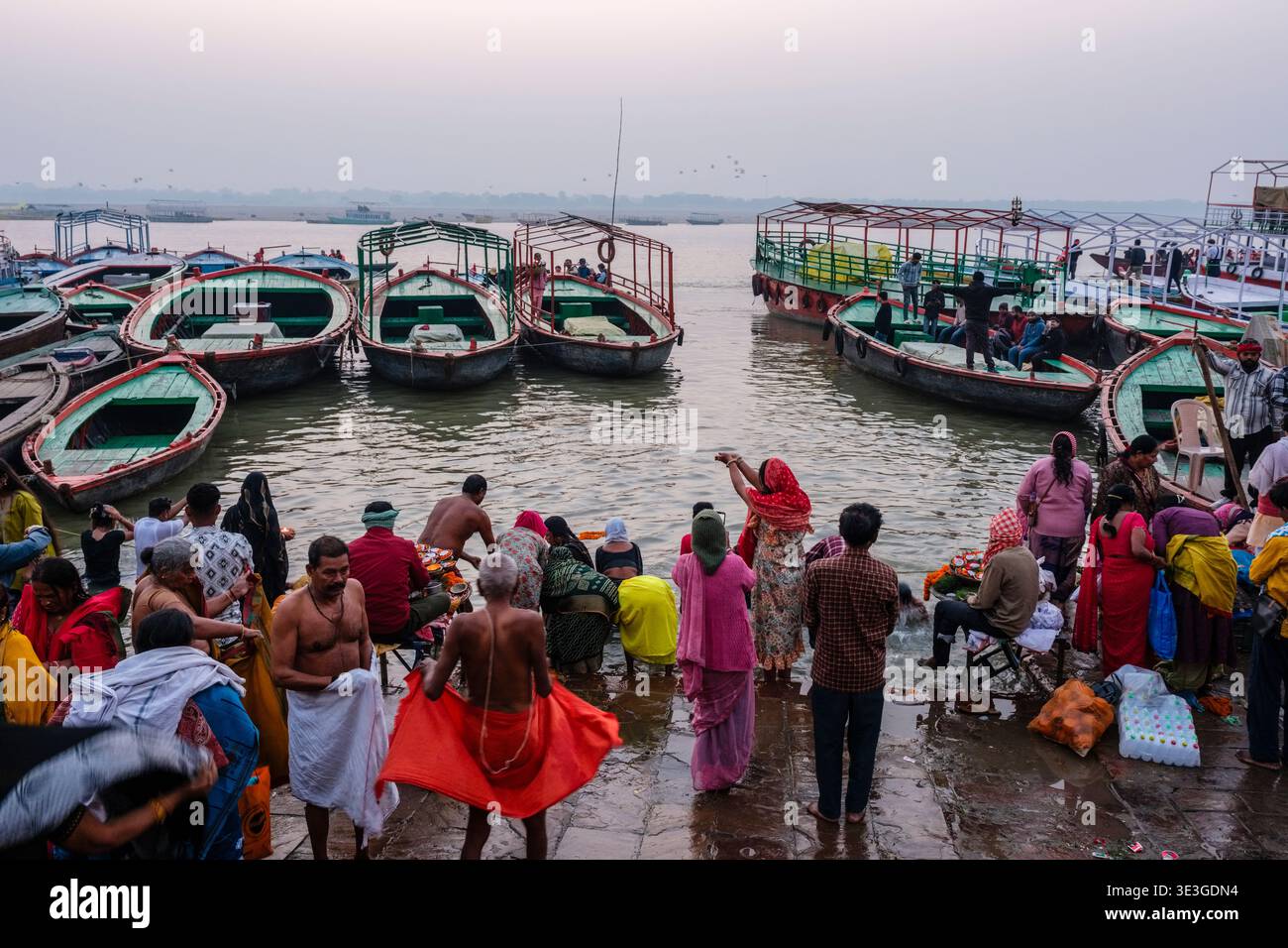 Des foules de pèlerins et de visiteurs se rassemblent au bord du Gange à Varanasi où de nombreux bateaux en bois vert et rouge sont amarrés à l'embarcadère. Banque D'Images