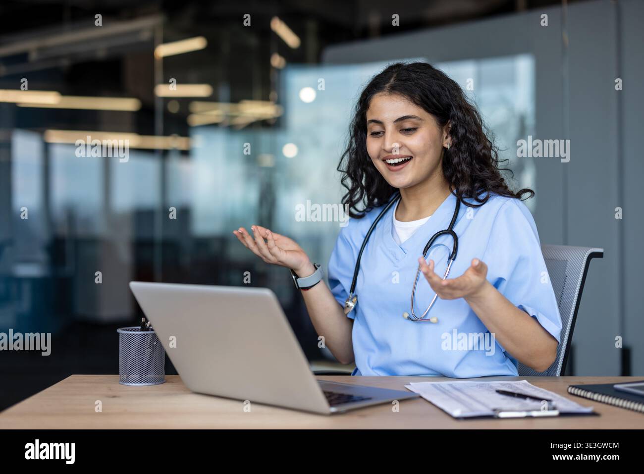 Jeune femme indienne médecin dans les gommages et stéthoscope souriant et gestuel tout en ayant un appel vidéo sur son ordinateur portable, fournissant une consultation médicale en ligne à partir d'un bureau moderne Banque D'Images