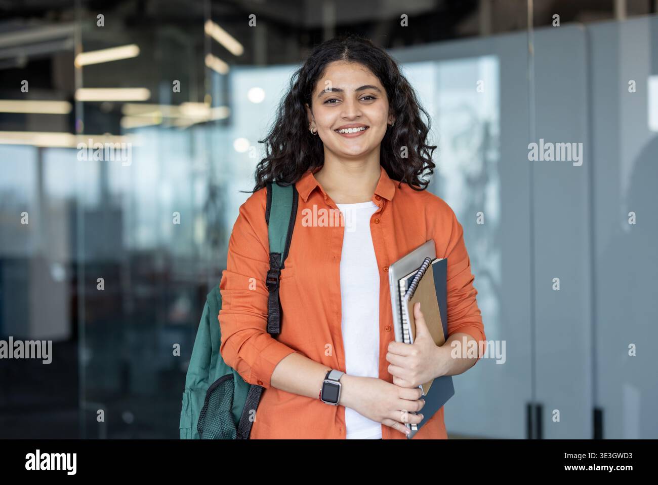Jeune étudiante indienne portant un sac à dos et des cahiers, debout dans une université moderne ou un immeuble de bureaux, regardant la caméra avec une expression confiante et heureuse Banque D'Images
