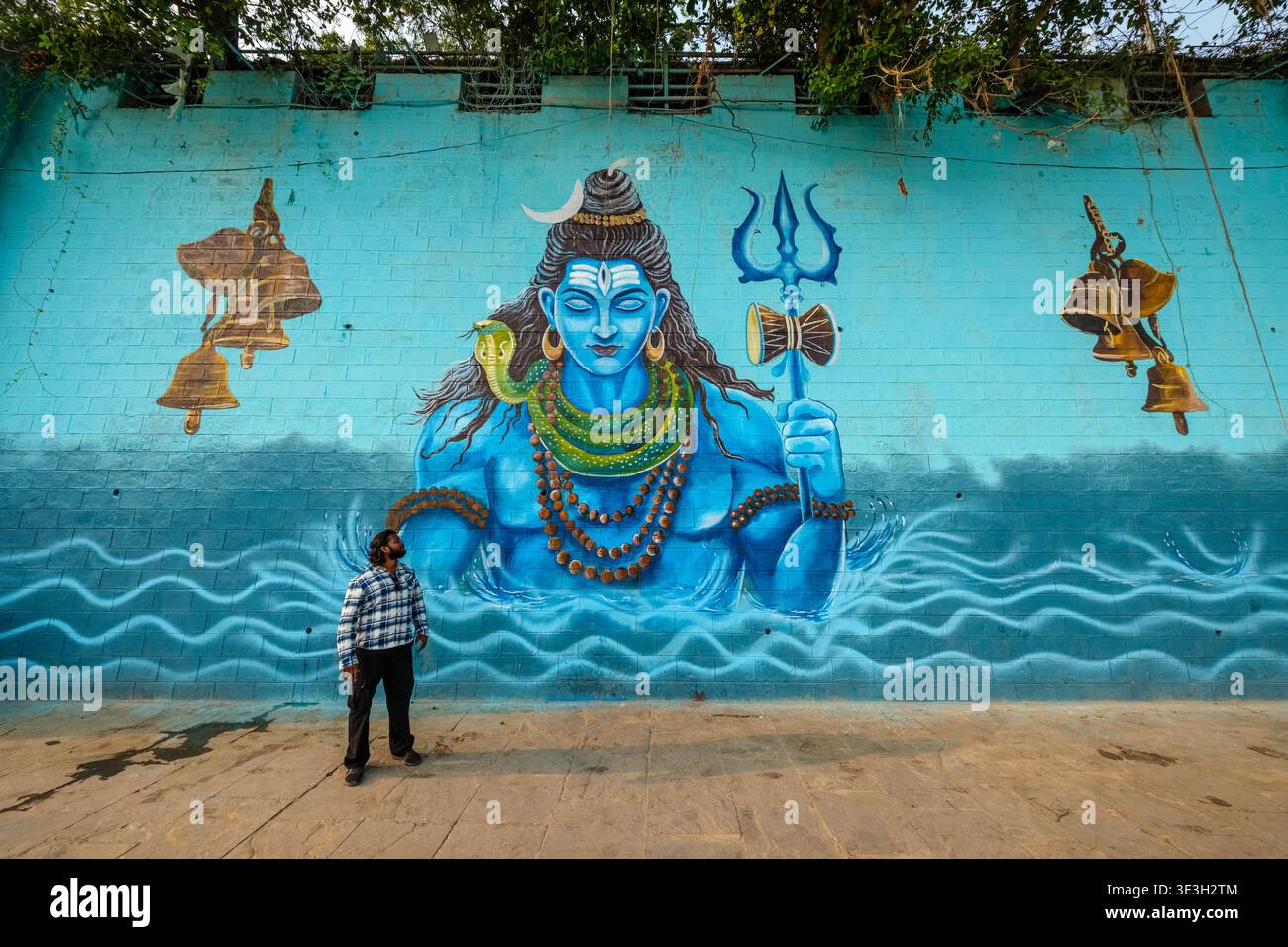 Un homme se tient devant une grande murale bleu vif de Lord Shiva peinte sur un grand mur dans un ghat Varanasi près du Gange. Banque D'Images