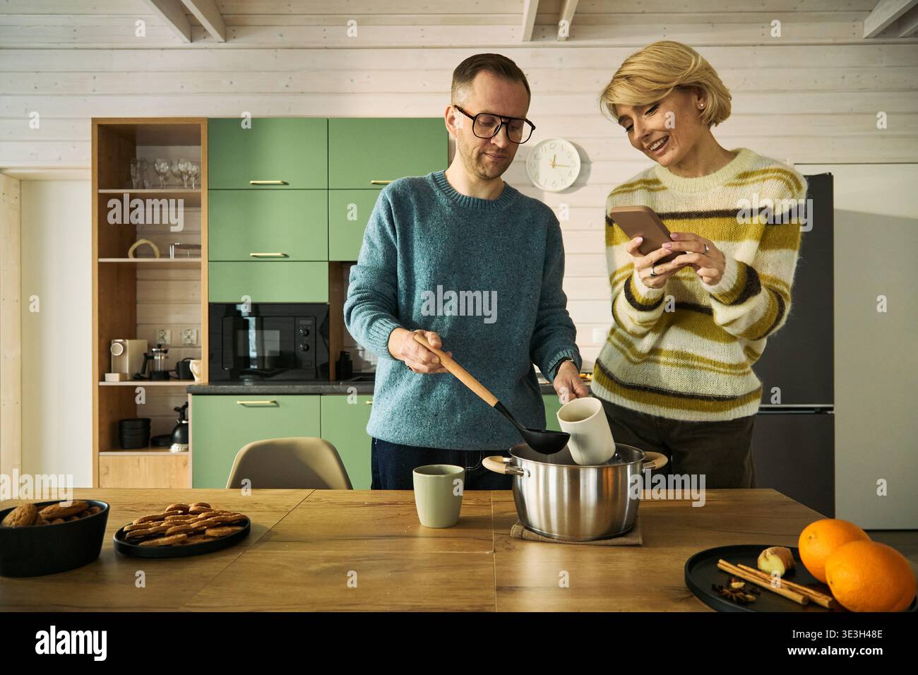 Homme d'âge moyen caucasien remuant le pot sur le comptoir de la cuisine tandis que femme d'âge moyen caucasienne debout à côté de lui tenant un smartphone et souriant à l'écran ensemble Banque D'Images