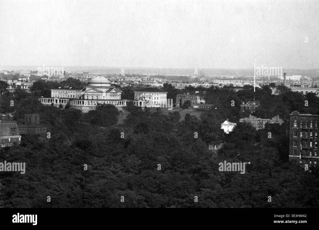 Cette photographie aérienne en noir et blanc, probablement des années 1920 ou 1930 par Angelo Rizzuto, capture une vue panoramique du Bronx Community College (alors probablement le campus University Heights de l'Université de New York) situé au sommet d'une colline dans le Bronx, New York. Les bâtiments néoclassiques, y compris la bibliothèque commémorative Gould en forme de dôme, sont en évidence parmi la couverture d'arbres luxuriante, avec le paysage urbain plus large visible au loin sous un ciel brumeux. Il offre un aperçu du développement architectural et urbain de New York à cette époque. Banque D'Images
