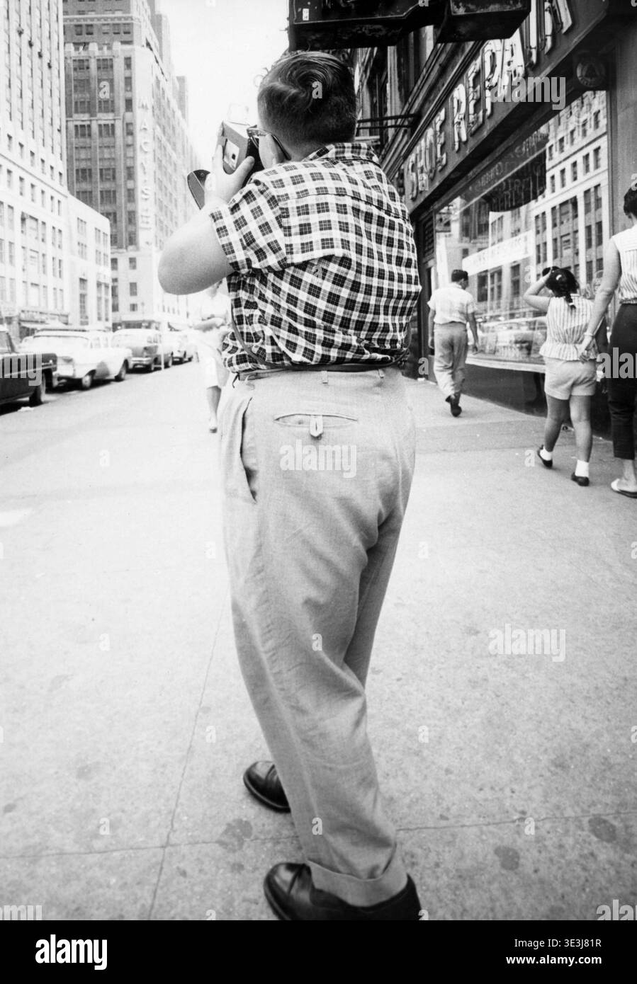 Capturée au milieu des années 1950 par le photographe Angelo Rizzuto, cette photographie de rue montre une personne par derrière, tenant un appareil photo et prenant apparemment une photo d'un paysage urbain animé de New York. La scène comprend des piétons et des voitures garées avec une vue d'un bâtiment affichant l'enseigne du grand magasin Macy's en arrière-plan. Banque D'Images
