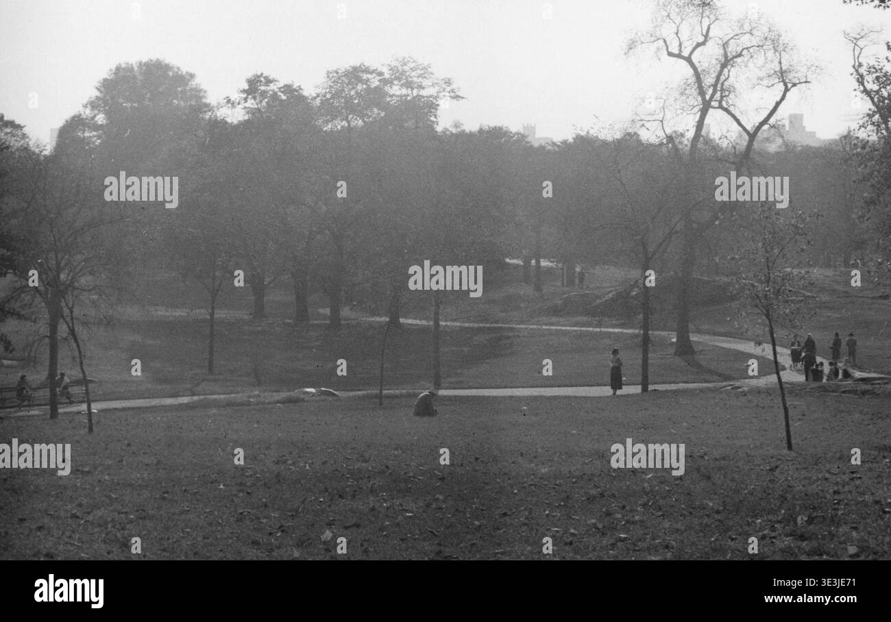 Une photographie tranquille en noir et blanc d'Angelo Rizzuto des années 1940 capture une vue panoramique de Central Park à New York. Les gens se promènent le long de sentiers sinueux et se détendent sur des bancs au milieu d'arbres luxuriants, avec des contours pâles des bâtiments de la ville visibles dans la distance brumeuse, offrant un aperçu de la vie quotidienne et de la mode de l'époque. Banque D'Images