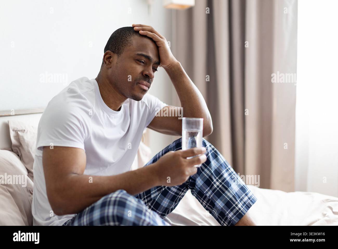 Homme noir assis sur le lit tenant un verre d'eau et touchant la tête Banque D'Images