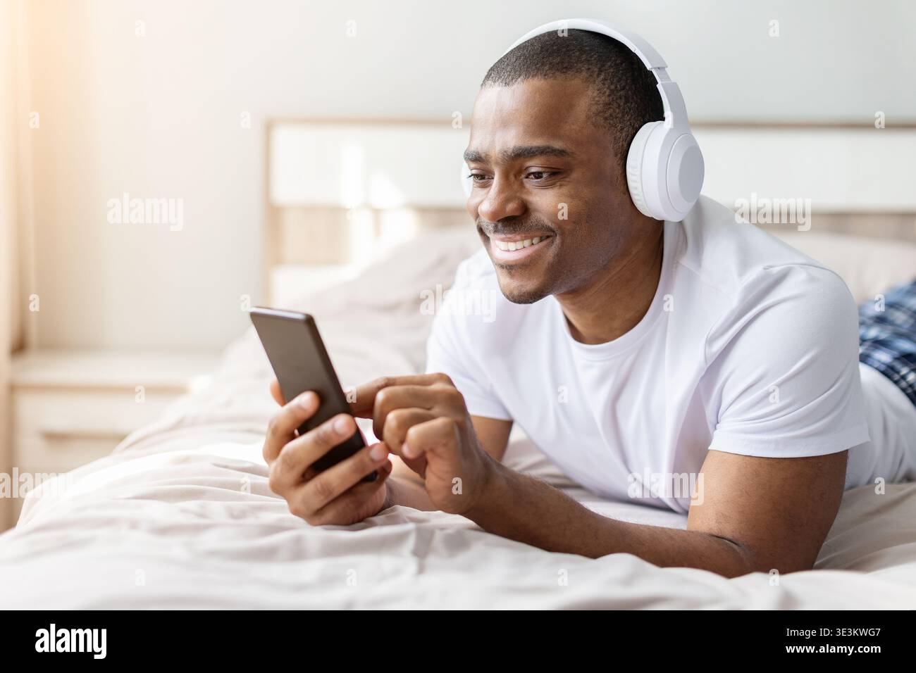Jeune homme afro-américain allongé sur le lit à l'aide d'un smartphone avec des écouteurs Banque D'Images