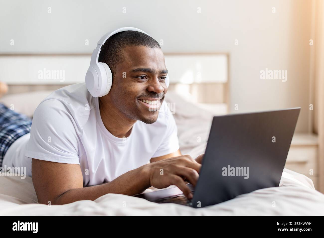 Jeune homme noir allongé sur le lit à l'aide d'un ordinateur portable et portant des écouteurs Banque D'Images