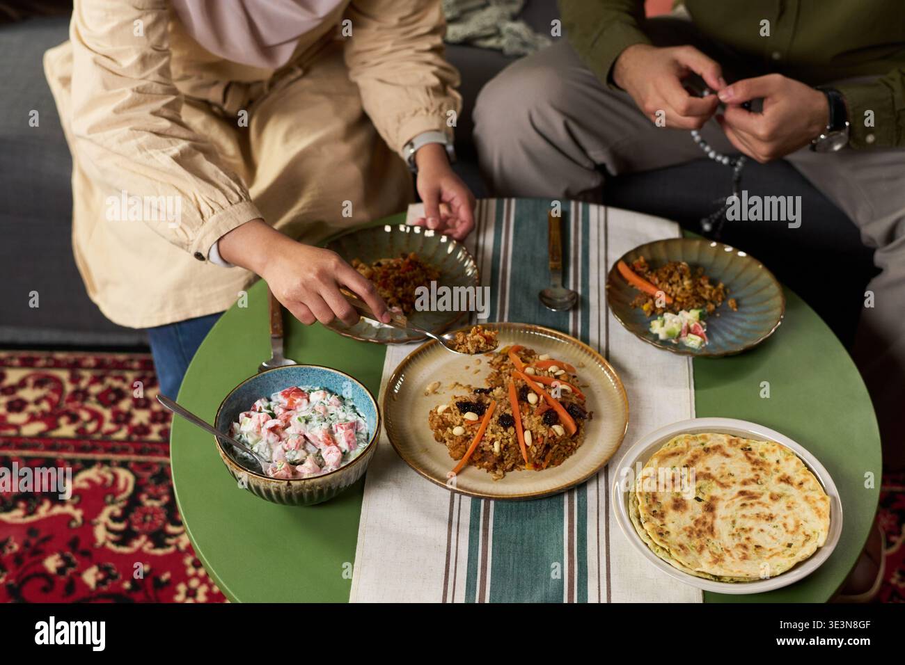 Femme musulmane et homme musulman assis à table partageant le repas traditionnel, femme atteignant pour la nourriture tandis que l'homme tenant des perles de prière, plats disposés sur la table verte Banque D'Images