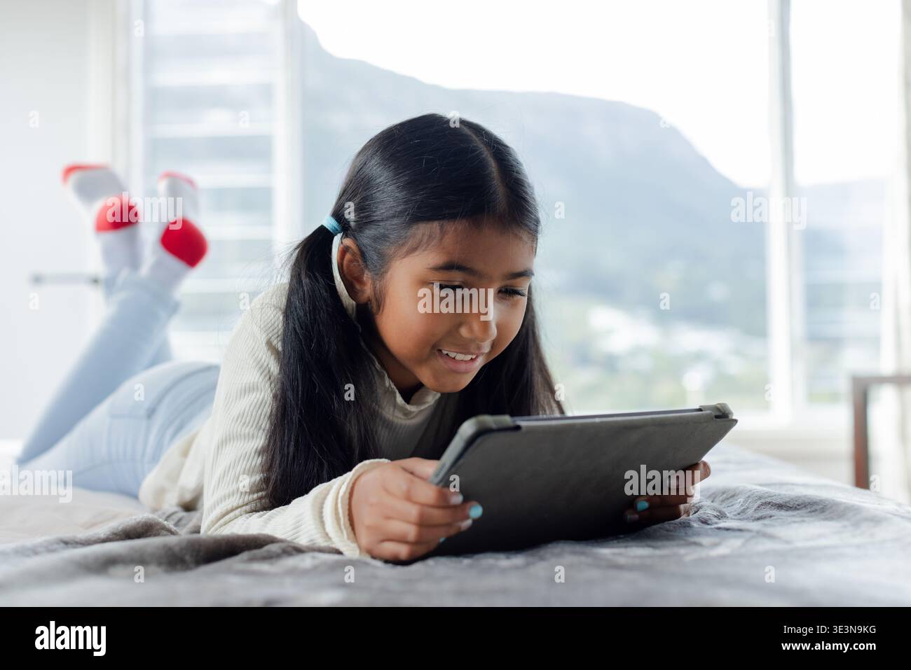 Fille d'âge scolaire utilisant une tablette noire alors qu'elle était allongée sur le lit avec une couverture grise dans la chambre près de la fenêtre Banque D'Images
