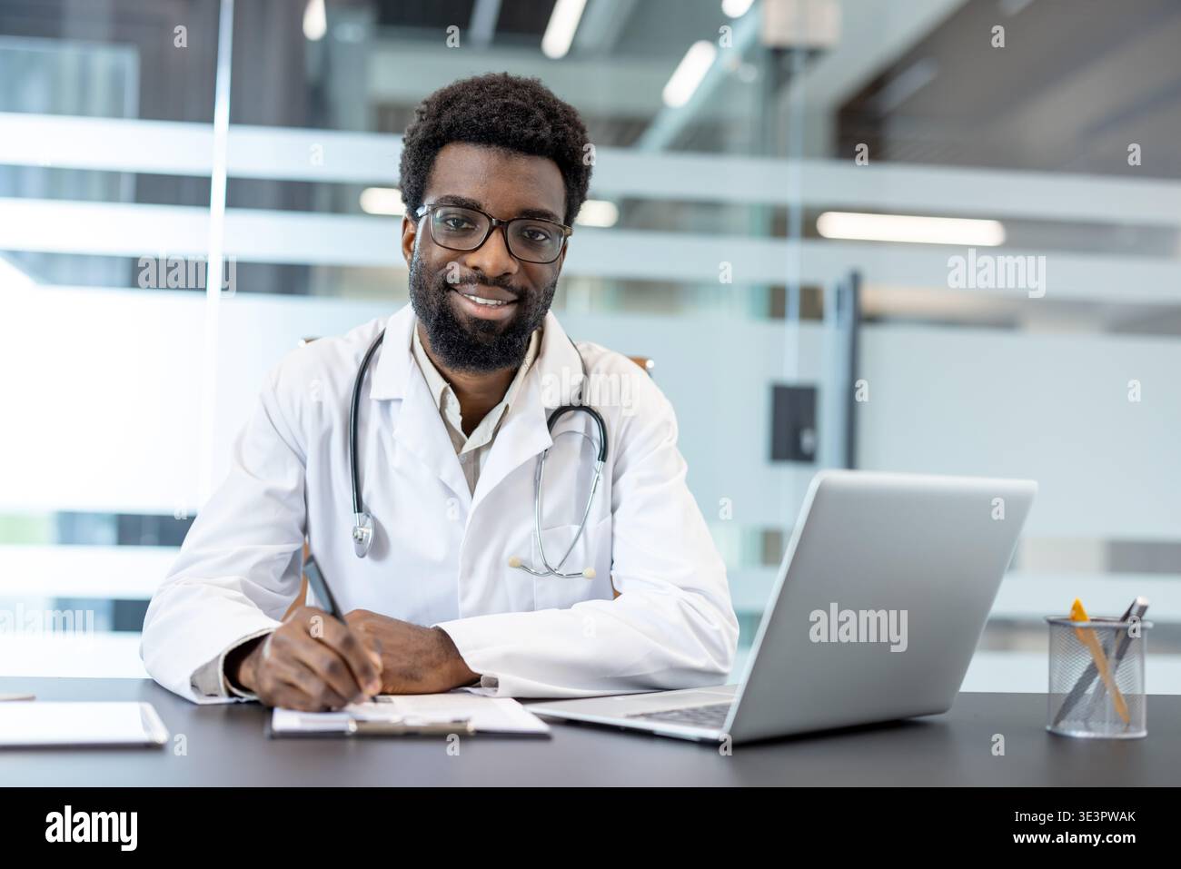 Médecin masculin confiant travaillant dans un bureau de clinique occupé, souriant et écrivant des notes tandis qu'un ordinateur portable et un stéthoscope sont visibles, représentant une guérison dédiée Banque D'Images