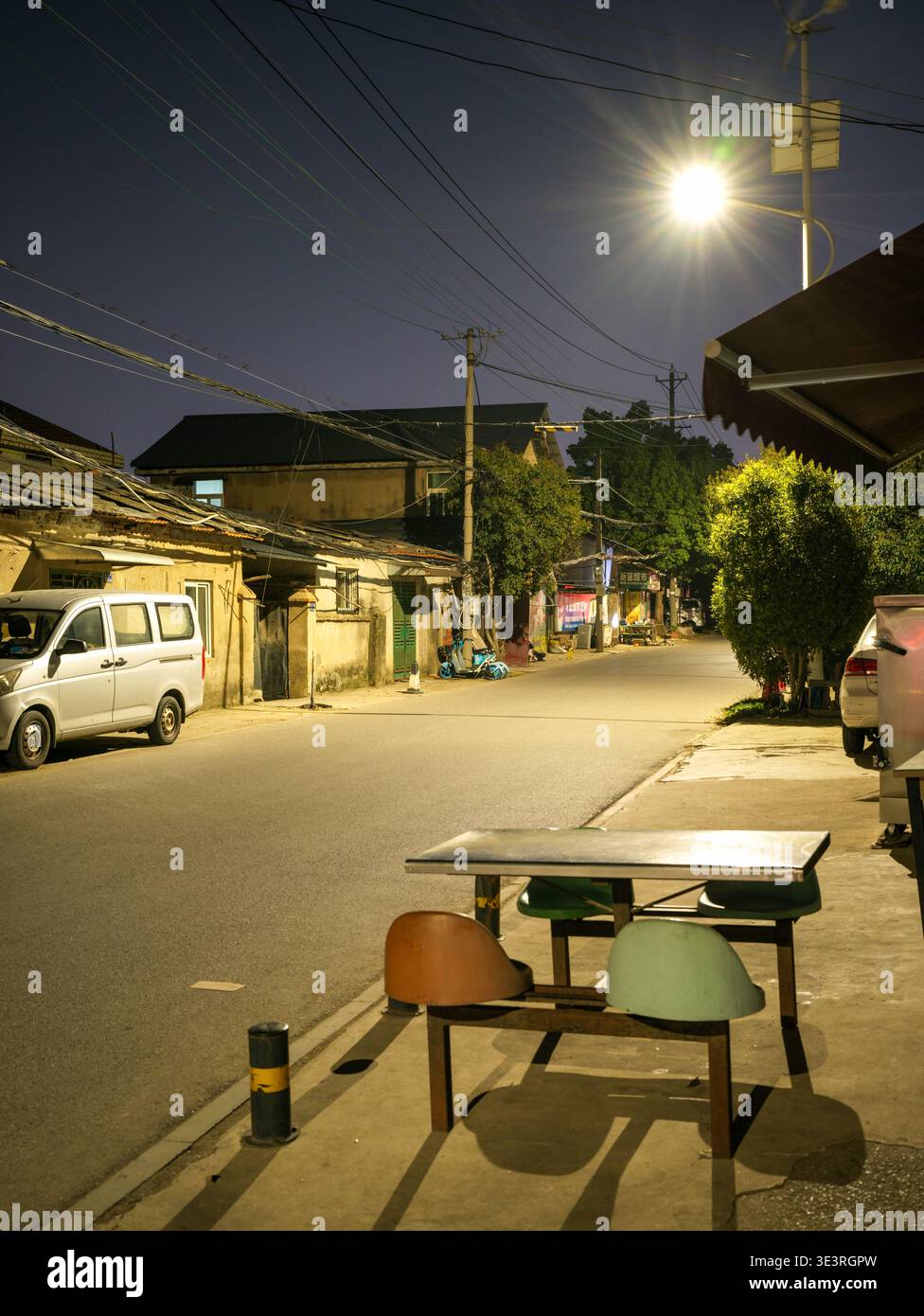 Une rue de village vide illuminée par un lampadaire lumineux révélant une petite table extérieure et des chaises dressées au bord de la route la nuit. La scène repr Banque D'Images