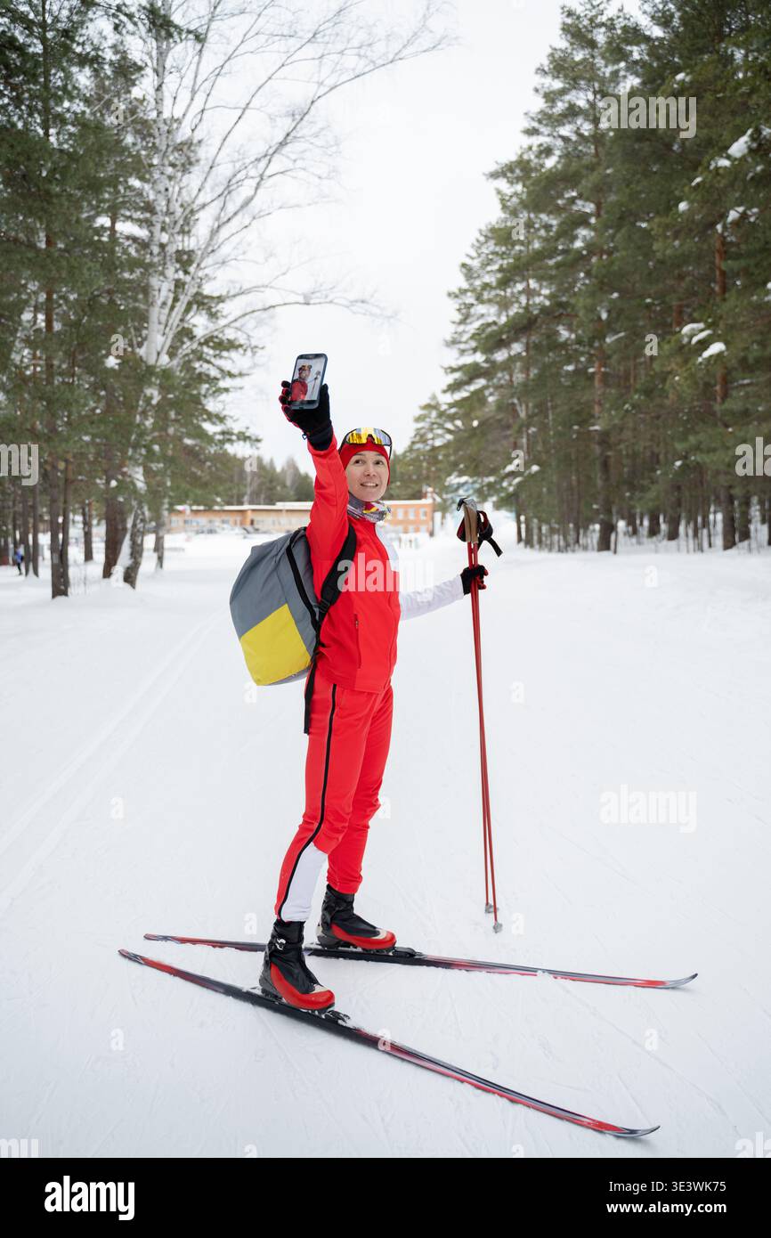 Un skieur de fond vêtu d'une tenue rouge vif prend un selfie au milieu d'une forêt enneigée en toile de fond. Le skieur tient des bâtons de ski, capturant la terre d'hiver sereine Banque D'Images