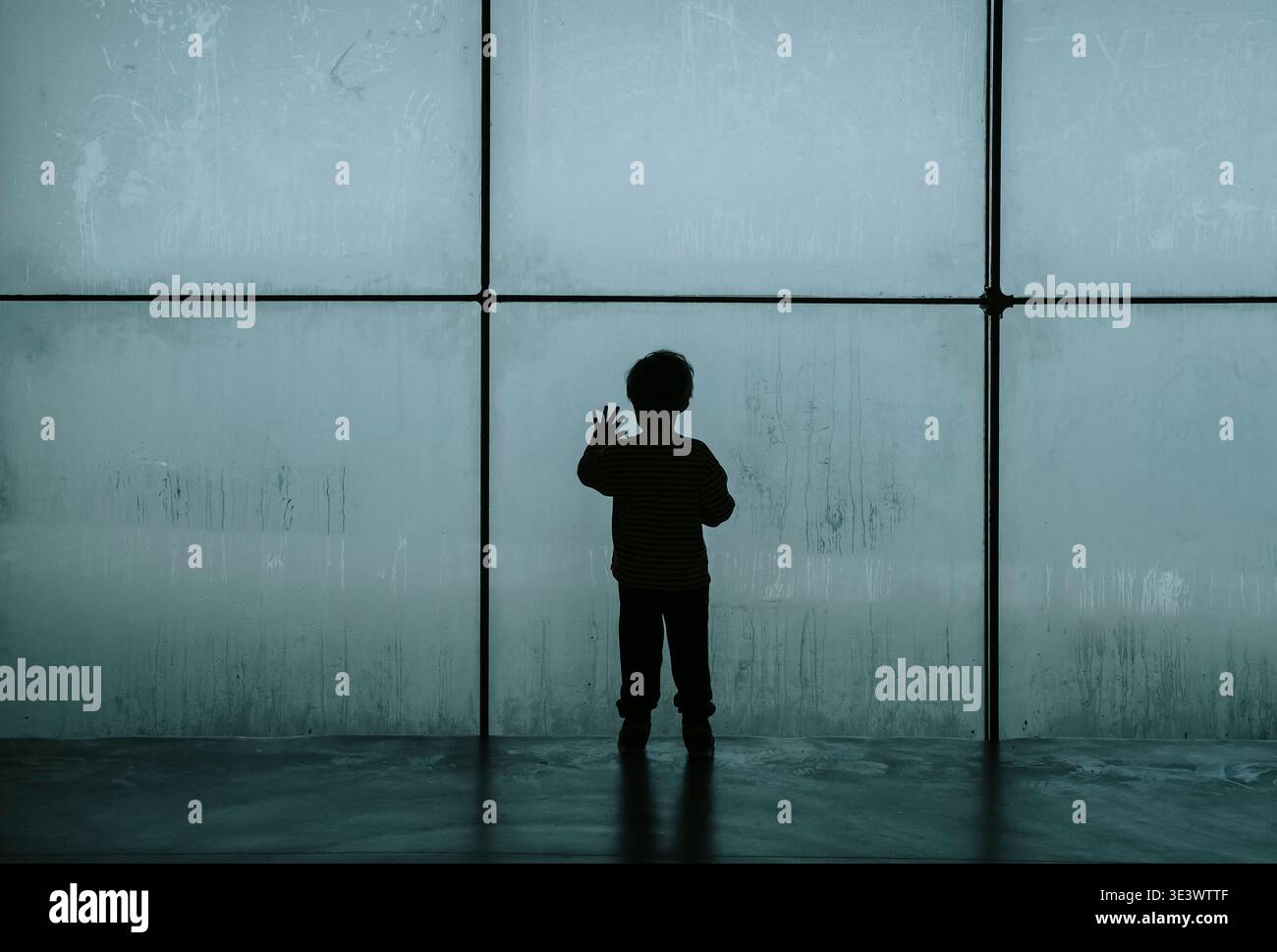 Silhouette d'un enfant solitaire regardant à travers une grande fenêtre embuée froide. Un travail de photographie conceptuelle explorant les thèmes de la dépression infantile, de la crise psychologique et du profond sentiment d’isolement et de tristesse. Photo de haute qualité Banque D'Images